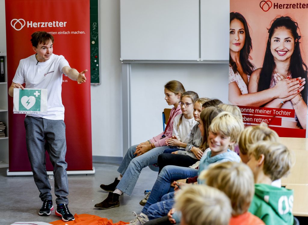HerzretterTrainer Julian klärt eine Gruppe Schüler:innen über den Defibrillator auf